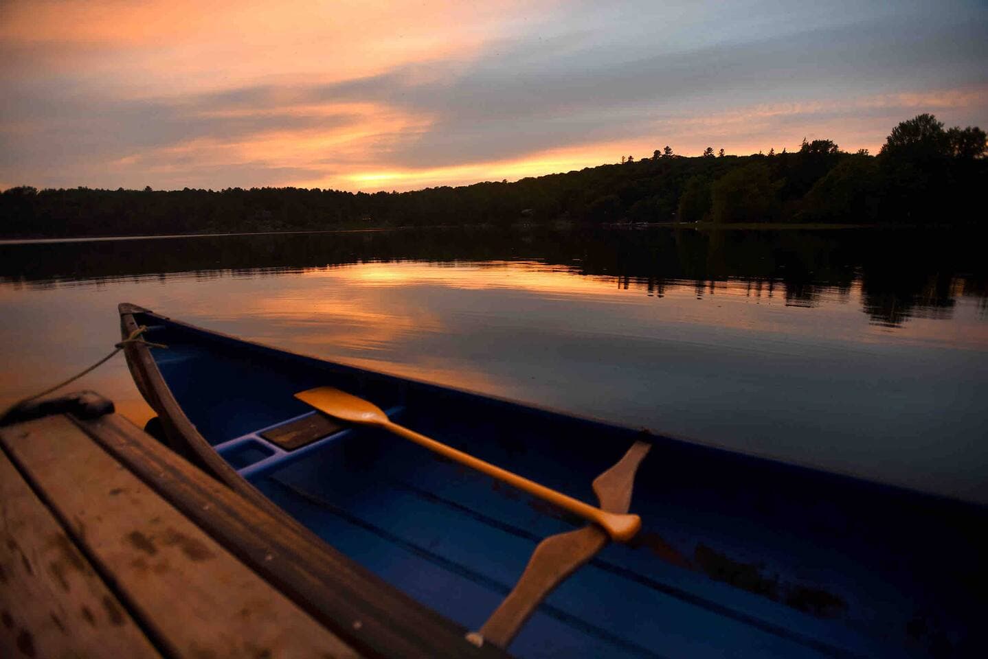 A blue canoe with wooden paddle rests at the dock during a glowing Three Mile Lake sunset.